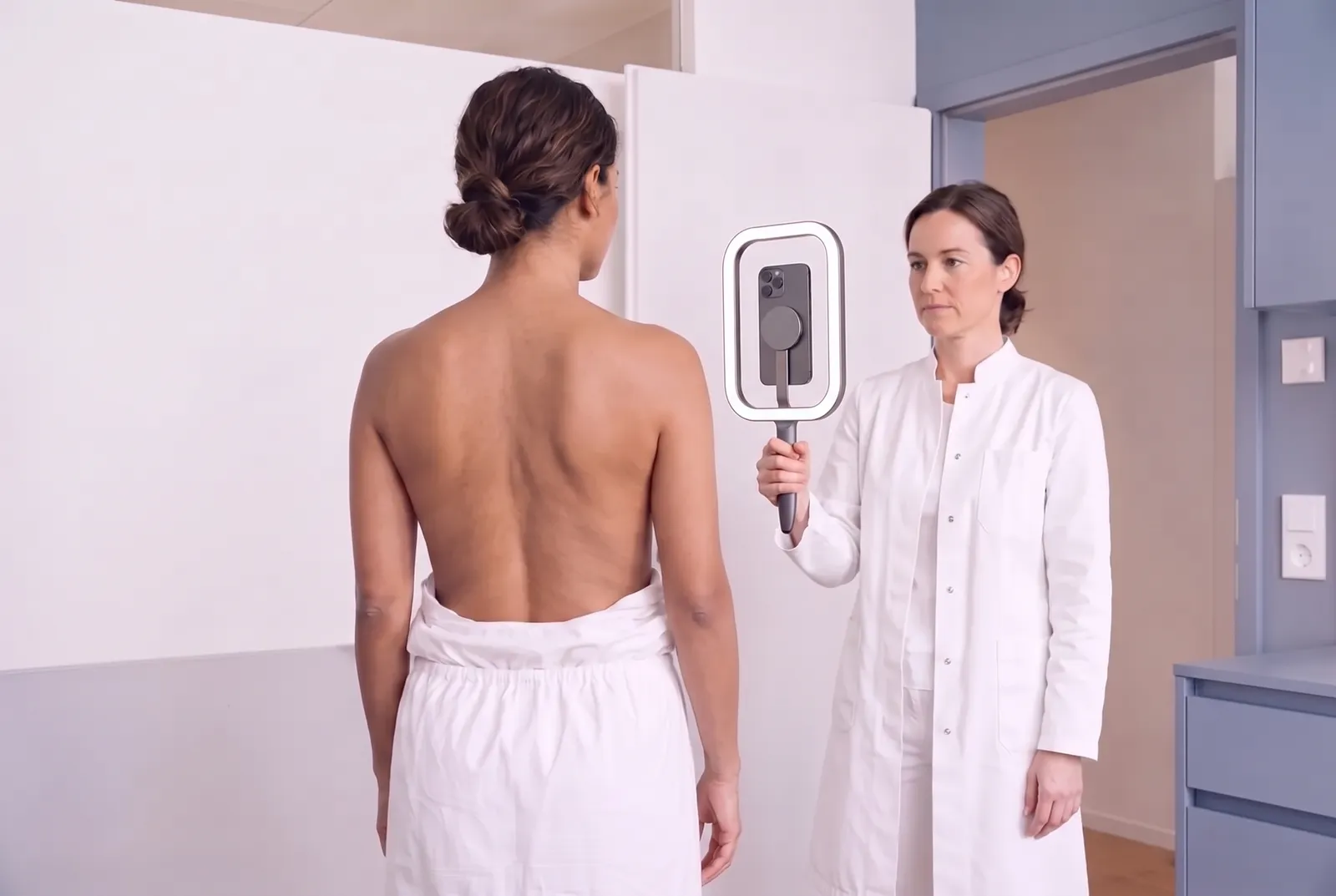 Eine Ärztin fotografiert den Oberkörper einer Patientin mit dem iPhone im Handlicht; die Patientin steht mit dem Rücken zum Betrachter, ein Handtuch um die Hüfte